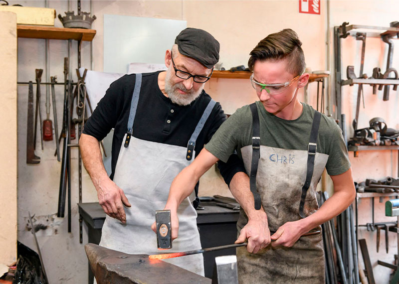 Foto: Schmied zeigt einem Jungen Hammer und glühendes Metall auf Amboss