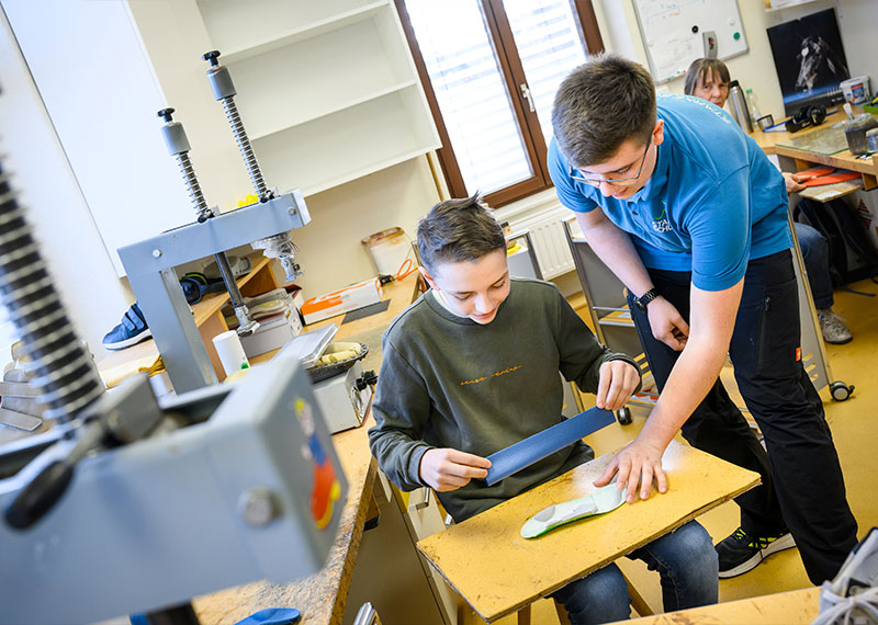 Foto: Ein Jugendlicher zeigt einem Jungen eine Schuhsohle in einer Werkstatt