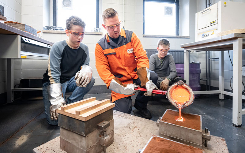 Foto: Ein Metallgießer zeigt zwei Jugendlichen das Metallgießen in eine Form