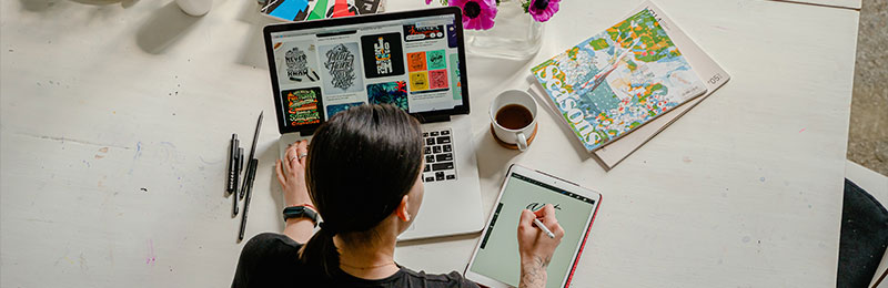 Foto: Eine Frau betrachtet Bilder auf einem Laptop, während sie auf einem Tablet zeichnet.