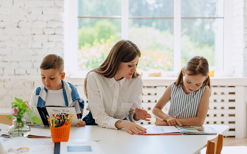 Foto: Ein Junge mit Buch sitzt neben einer Frau mit und einem Mädchen, welches ein Buch betrachtet. Auf dem stehen Tisch Blumen und bunte Schtifte.