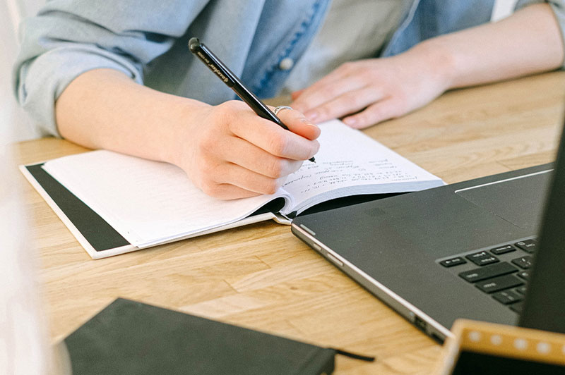 Foto: Hände mit Stift und ein Notizbuch neben einem Laptop
