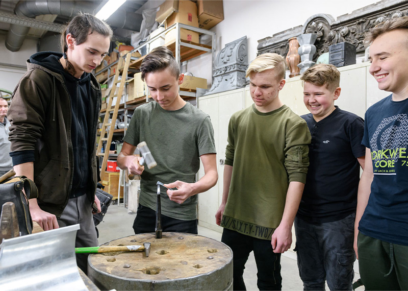 Foto: Vier Jungen schauen zu, wie ein weiterer Junge Metall mit dem Hammer bearbeitet.