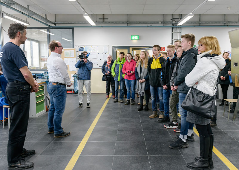 Foto: Zwei Männer und eine Person, die filmt, stehen vor Jugendlichen in einer Reihe.