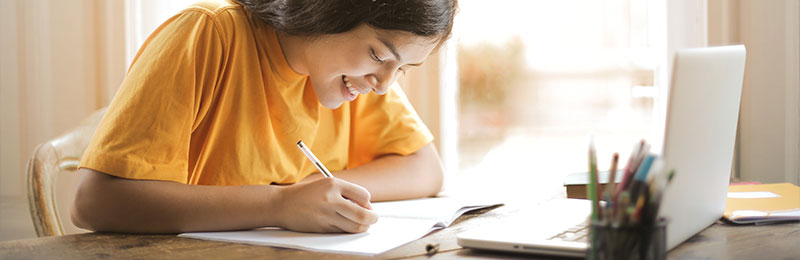 Foto: Junge schreibt im Notizbuch neben einem Fenster vor einem Laptop.