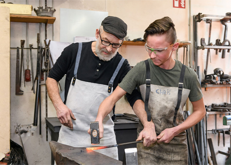 Foto: Ein Schmied zeigt einem Jungen Hammer und glühendes Metall auf einem Amboss.