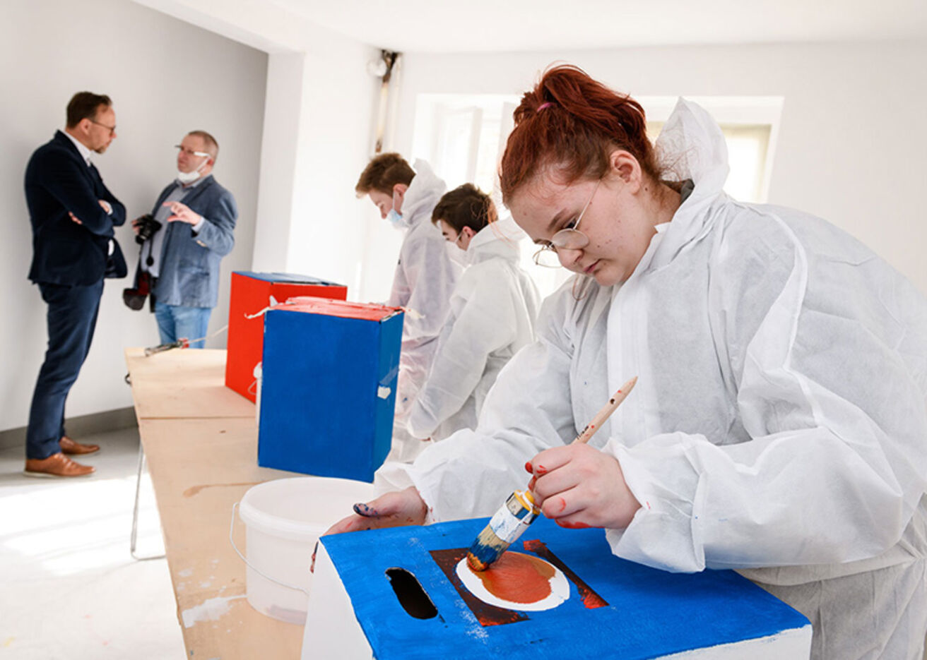 Foto: Drei Personen in Schutzanzügen bemalen Gegenstände mit Farbe. Zwei unterhalten sich im Hintergrund.