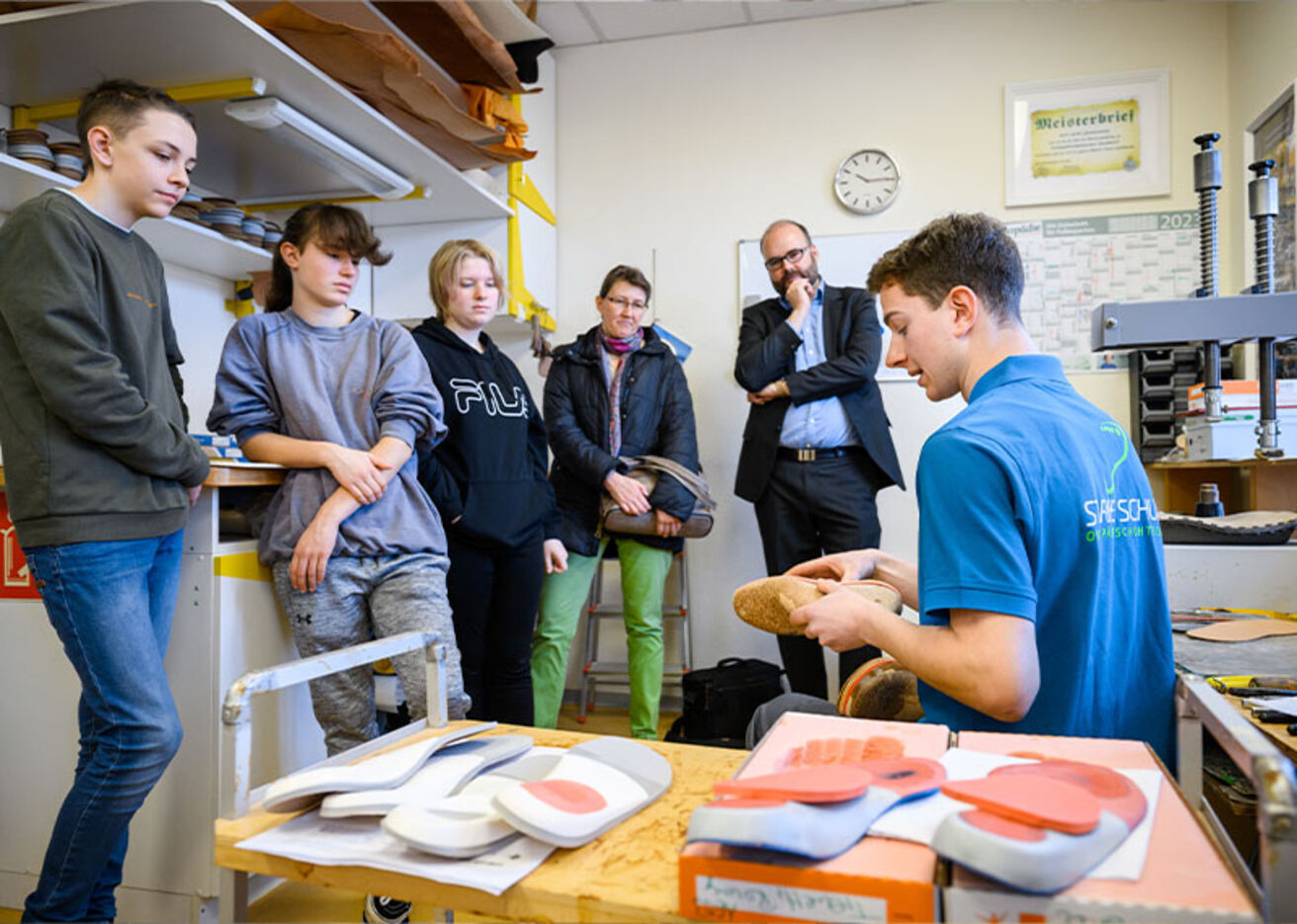 Foto: Jugendliche und Erwachsene schauen auf einen Jungen, welcher etwas zu dem Schuh in seiner Hand erklärt.