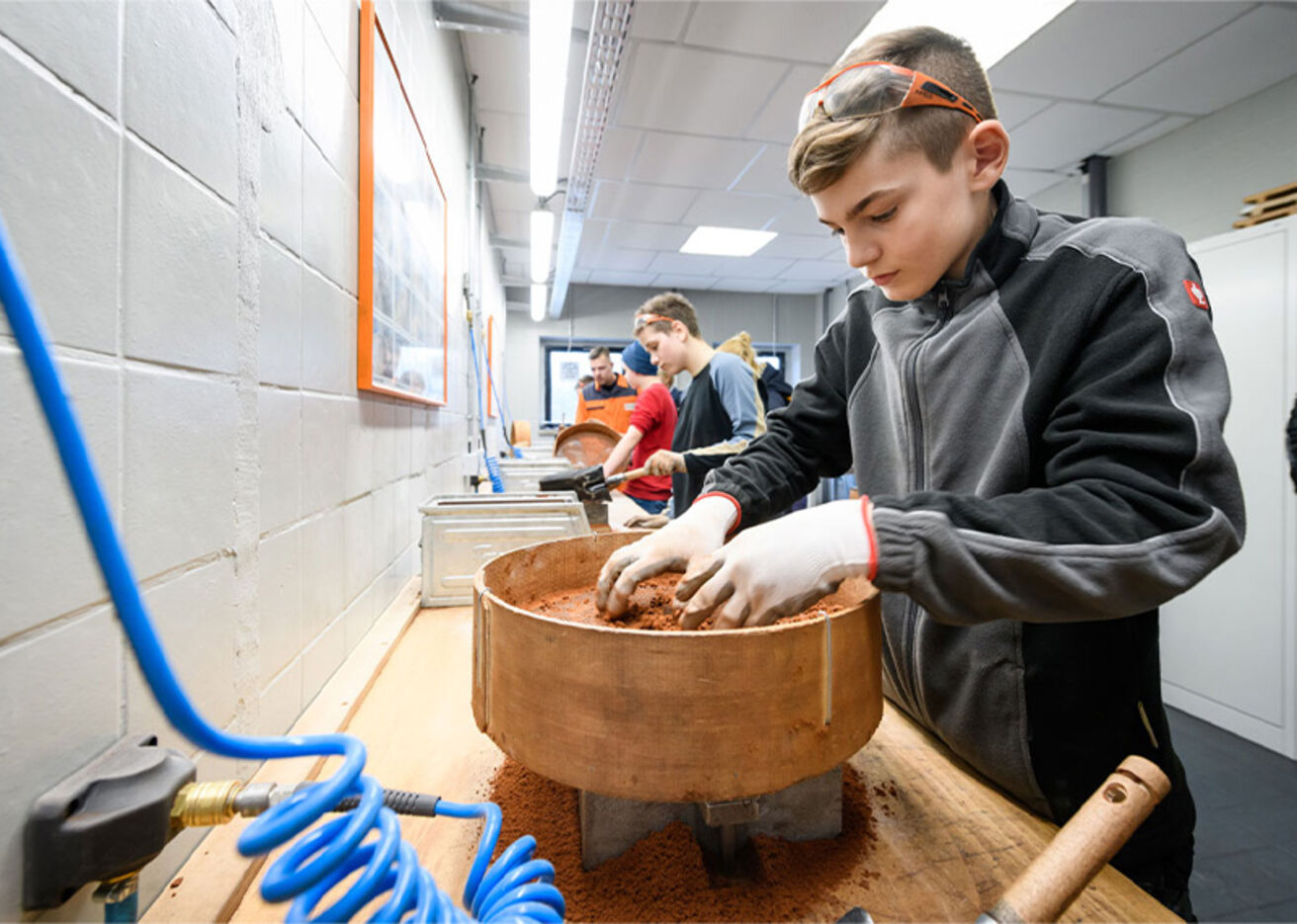 Foto: Ein Junge mit Handschuhen rührt in einem sandigen Material im Behälter.
