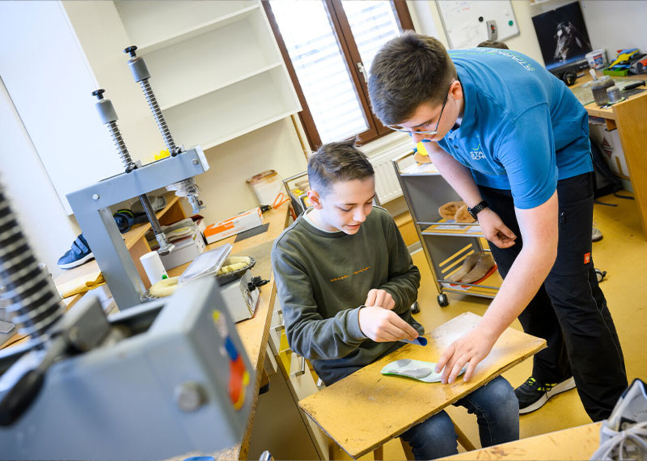 Foto: Ein Jugendlicher zeigt einem Jungen eine Schuhsohle in einer Werkstatt
