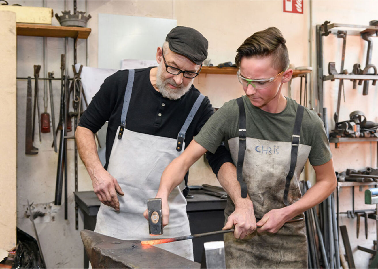 Foto: Schmied zeigt einem Jungen Hammer und glühendes Metall auf Amboss