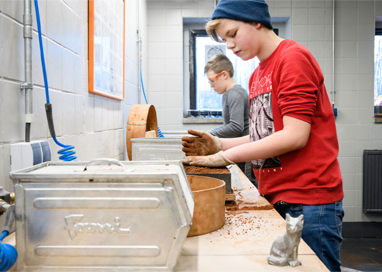 Foto: Ein Junge mischt eine rötlich-braune Substanz. Ein Schlauch steckt in einem Gerät und eine Skulptur steht auf dem Tisch. Ein weiterer Junge befindet sich im Hintergrund.